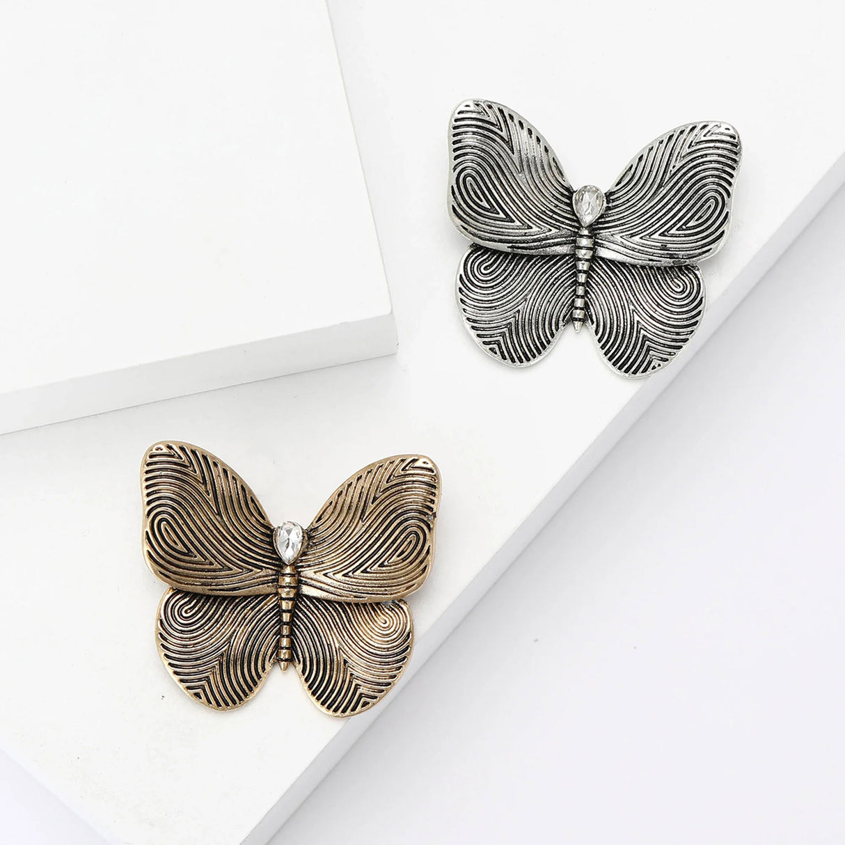 Gold-tone and silver-tone butterfly brooches with swirl wing etching and clear teardrop stones, flat lay on white.