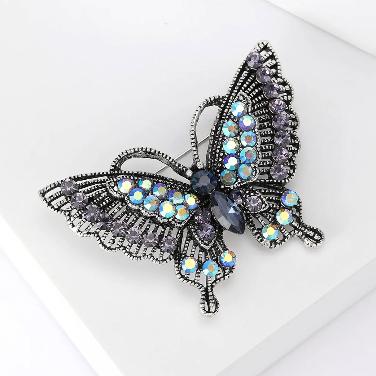 Butterfly brooch with dark silver-tone metal, iridescent black and blue stones, angled view on white studio backdrop.