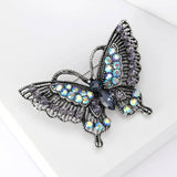 Butterfly brooch with dark silver-tone metal, iridescent black and blue stones, angled view on white studio backdrop.