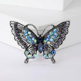 Butterfly brooch with dark silver-tone metal finish, blue faceted center, iridescent black-blue accent stones, front view on white block.