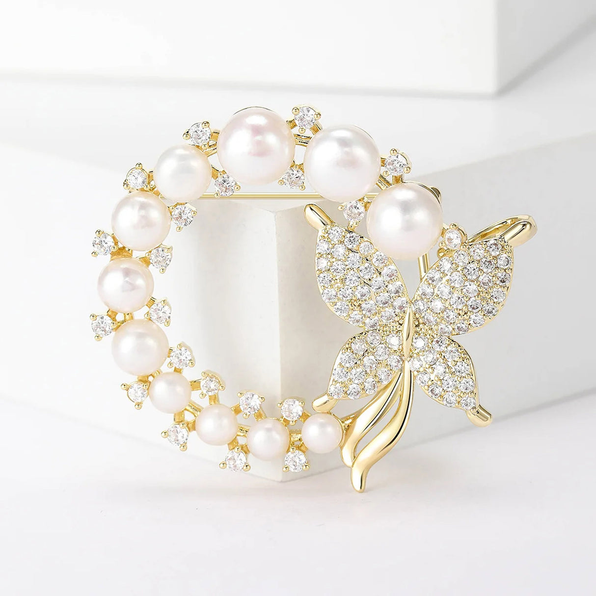 Butterfly brooch in gold-tone metal with white round beads and clustered clear stones, standing on white block.