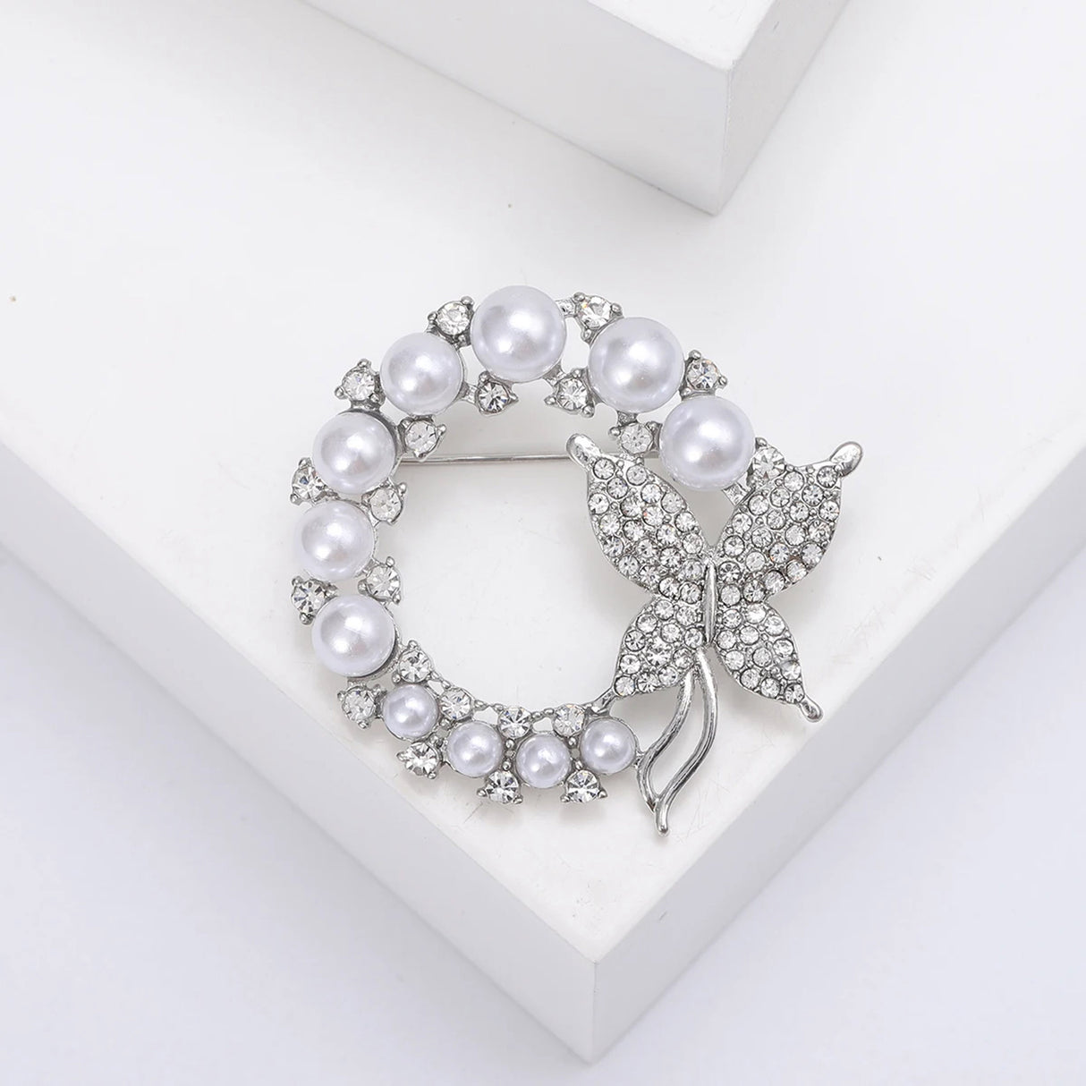 Butterfly brooch with silver-tone metal, glossy white round beads, and clear faceted stones, flat lay on white block.