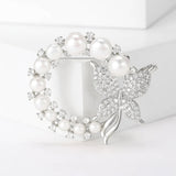 Silver-tone butterfly brooch with round white imitation pearls and clustered clear stones in circular layout, standing on white cube.