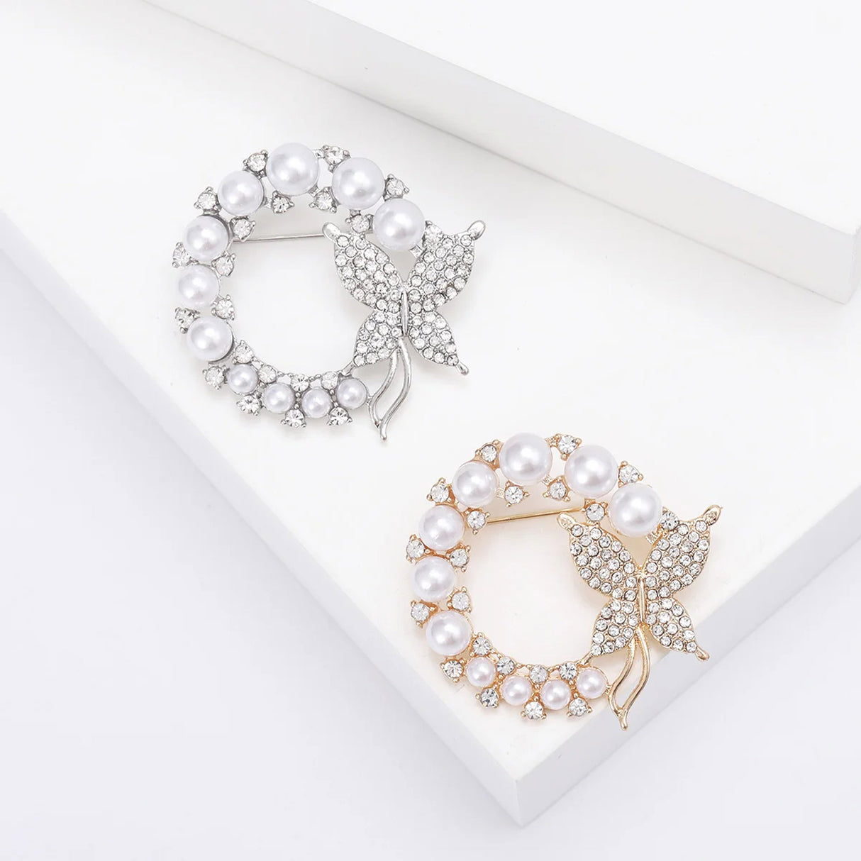 Set of two butterfly brooches in gold-tone and silver-tone metal with white round faux pearls and clear stones, flat lay on white surface.