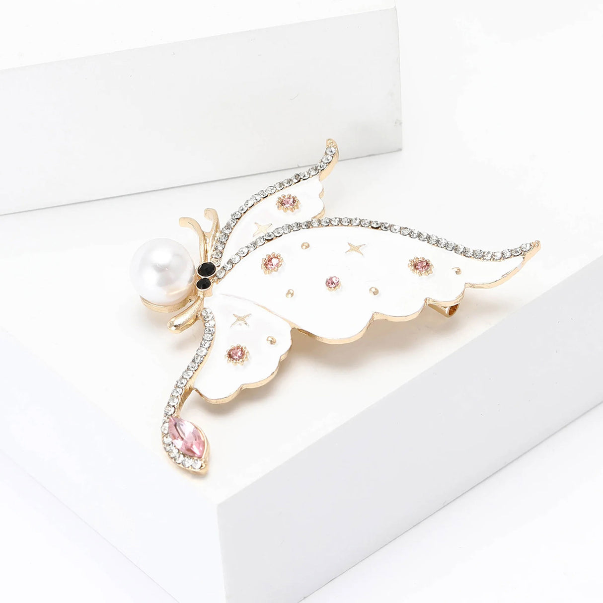 Butterfly brooch with white wings, pink round stones, pearl accent, and clear teardrop detail, angled view on white platform.