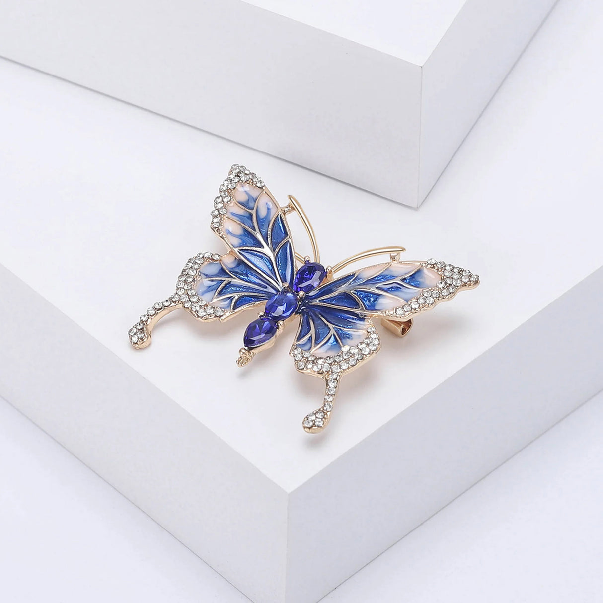Butterfly brooch with blue and white enamel wings, gold-tone frame, clear wing stones, and three faceted blue oval stones forming the body, angled view on white block.