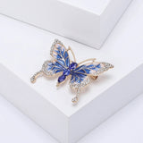 Butterfly brooch with blue and white enamel wings, gold-tone frame, clear wing stones, and three faceted blue oval stones forming the body, angled view on white block.