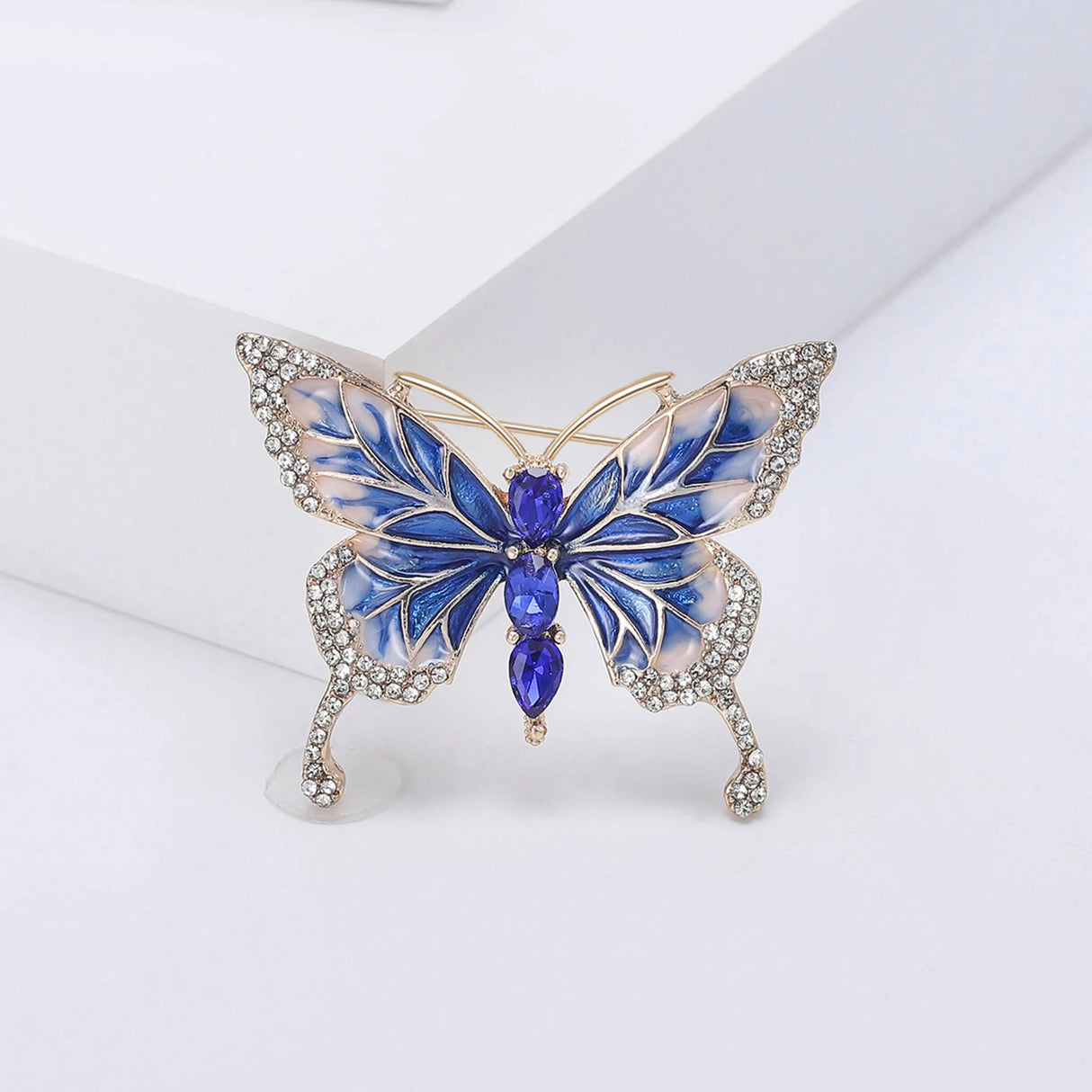 Butterfly brooch with deep blue and white enamel wings, gold-tone outline, clear decorative stones, and vertical row of blue faceted oval stones at center, upright pose on white.