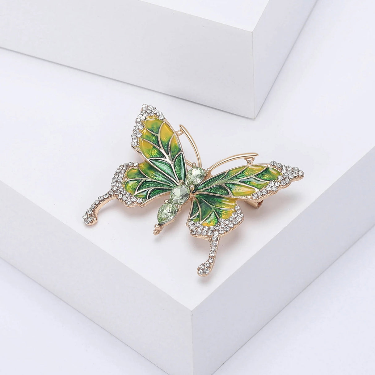 Butterfly brooch with green and yellow enamel wings, gold-tone edging, clear accent stones, and three green faceted ovals forming the body, angled view on white platform.