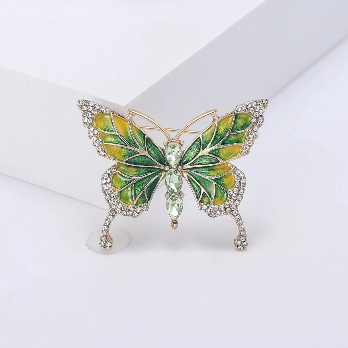 Butterfly brooch featuring green and yellow enamel wings, gold-tone trim, clear wing stones, and three green faceted oval stones at center, upright pose on white block.