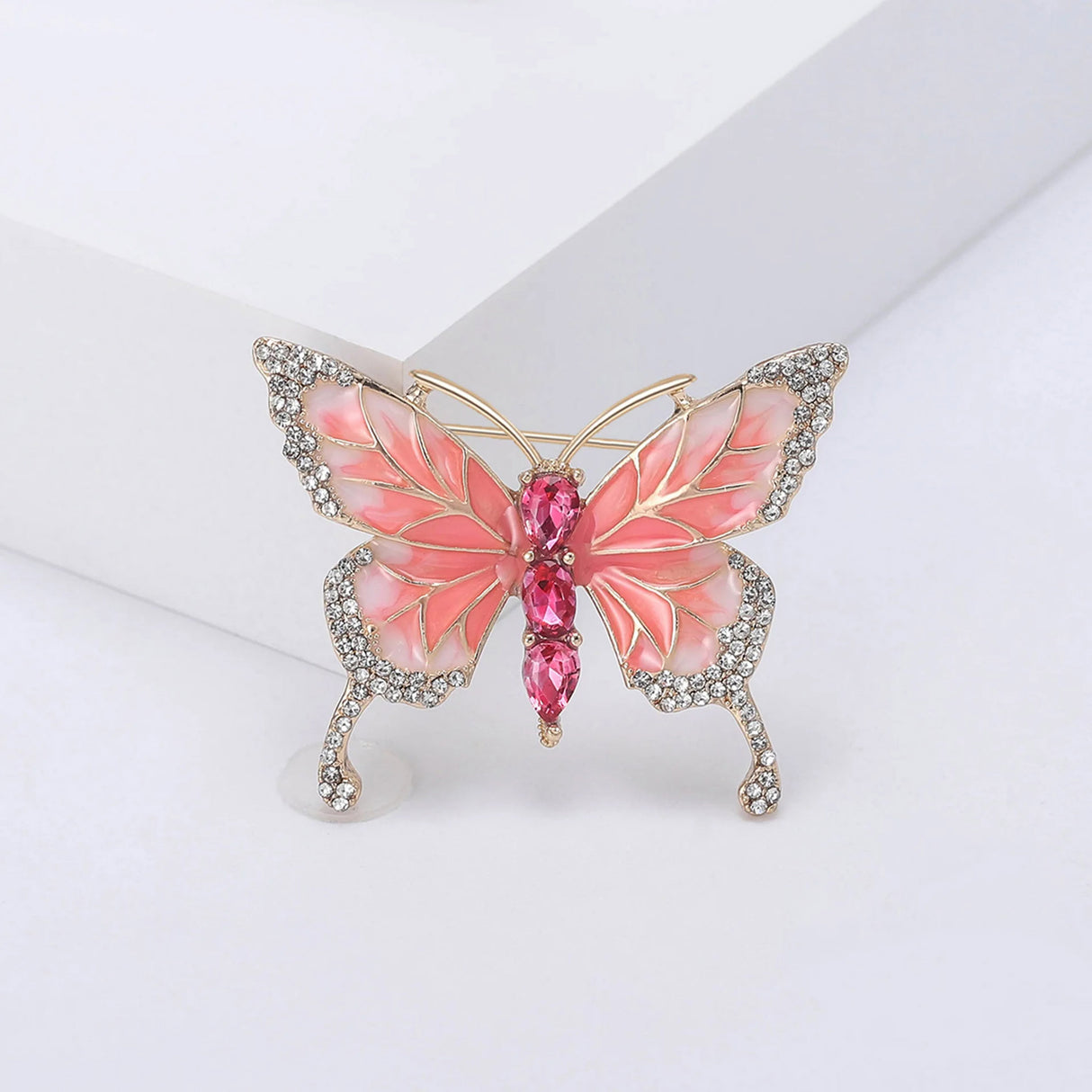 Butterfly brooch with coral-pink enamel wings, gold-tone metal, white small round stones along edges, and pink faceted oval stones forming the body. Upright pose on white studio background.
