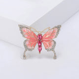 Butterfly brooch with coral-pink enamel wings, gold-tone metal, white small round stones along edges, and pink faceted oval stones forming the body. Upright pose on white studio background.