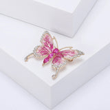 Butterfly brooch with pink enamel wings, gold-tone outline, clear edge stones, and three large vivid pink faceted ovals in the body, angled pose on white platform.
