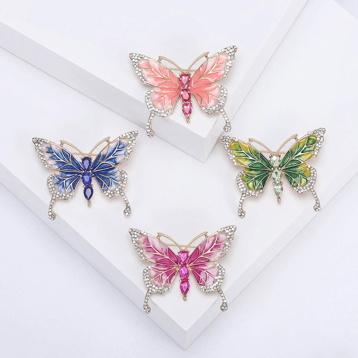 Set of four butterfly brooches with enamel wings in pink, green, and blue tones, each with small clear accent stones and large faceted oval body stones. Top-down view on white platforms.