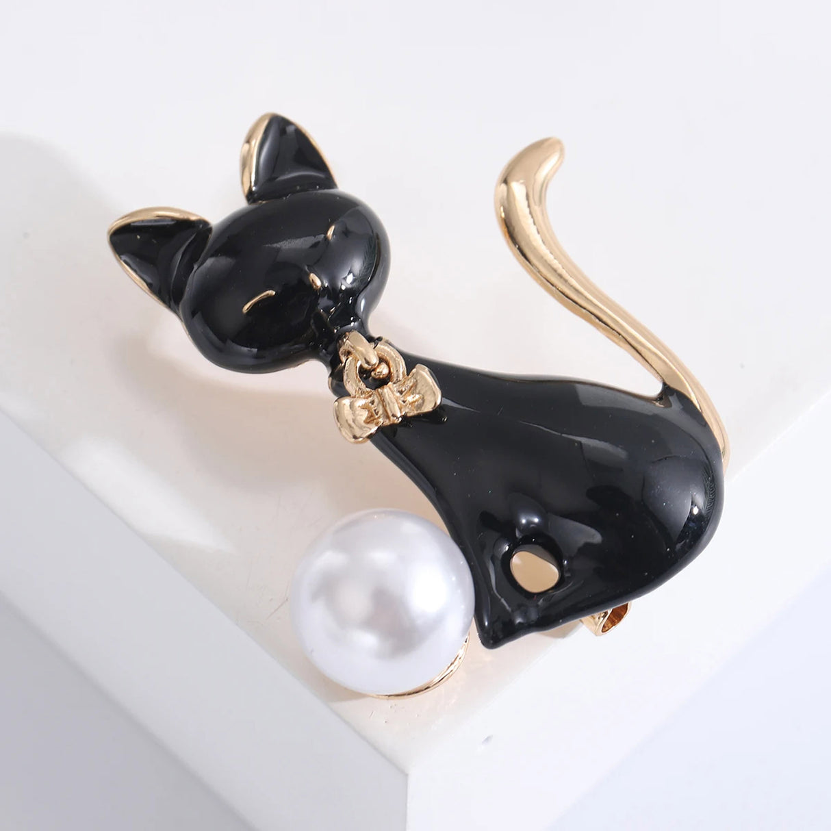 Cat brooch in black enamel with gold-tone trim and bow, paired with a white round bead, angled view on white studio backdrop.