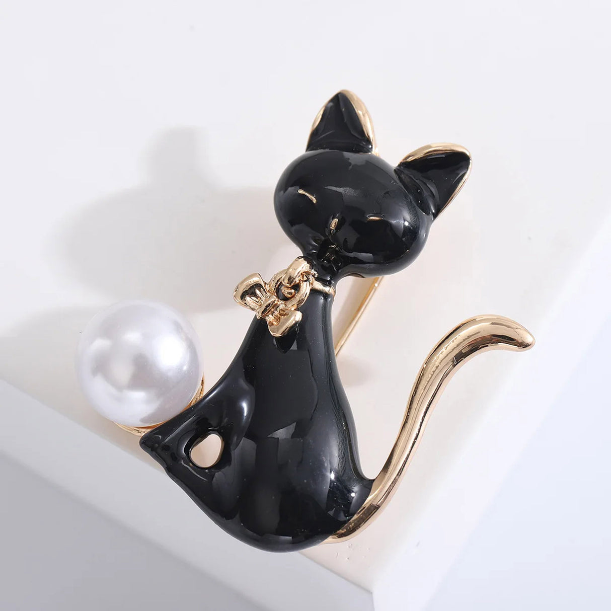 Cat brooch with black enamel finish, gold-tone tail and ears, and white round bead, angled top-down view on white studio surface.
