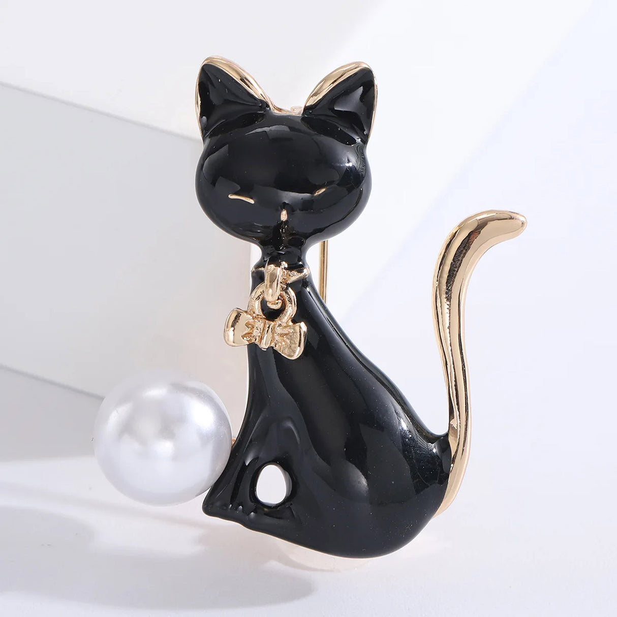 Cat brooch featuring black enamel body, gold-tone accents, and a white round bead, upright pose on white studio platform.