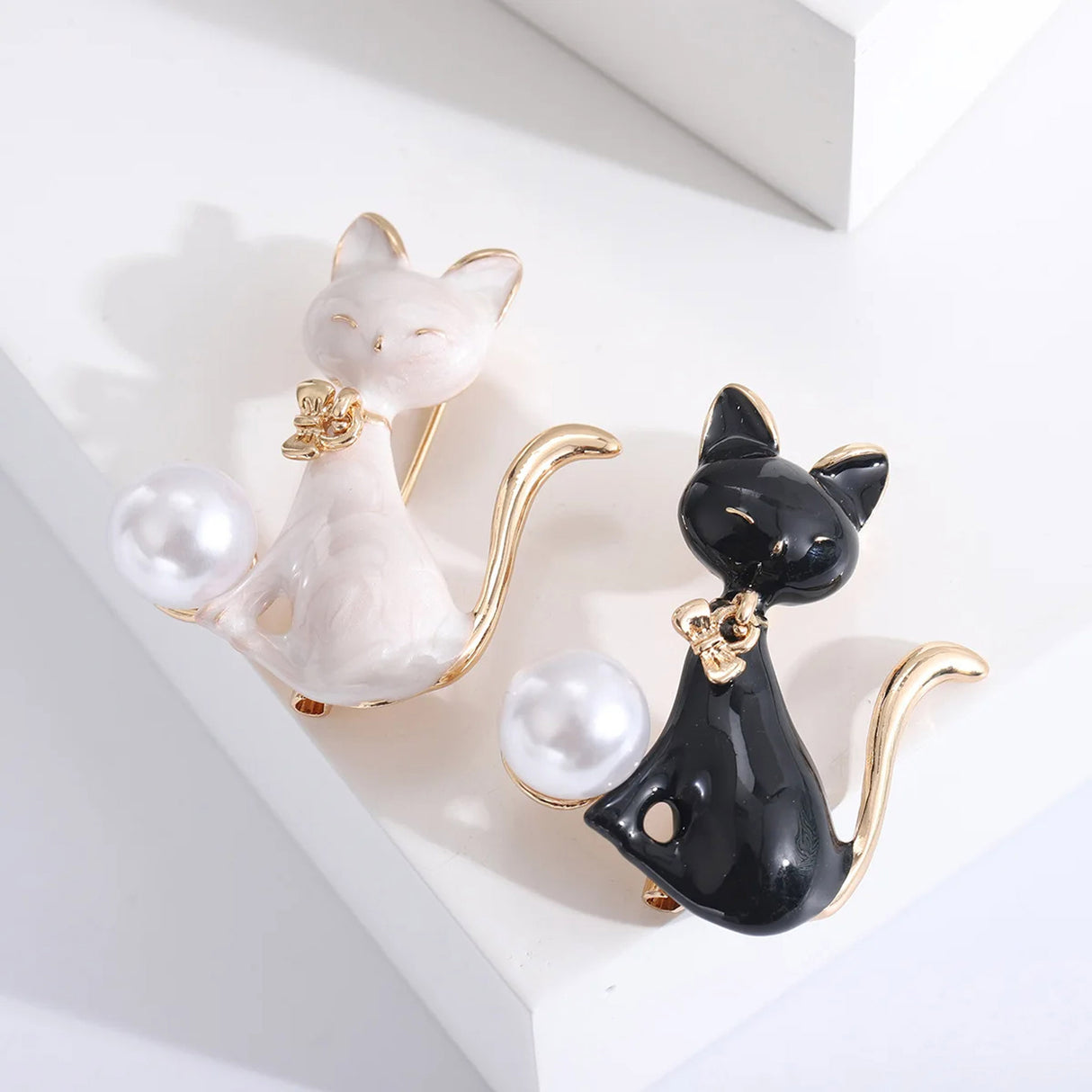 Set of two cat brooches, one in black enamel and one in white enamel, each with gold-tone trim and white round faux pearl, flat lay on white surface.