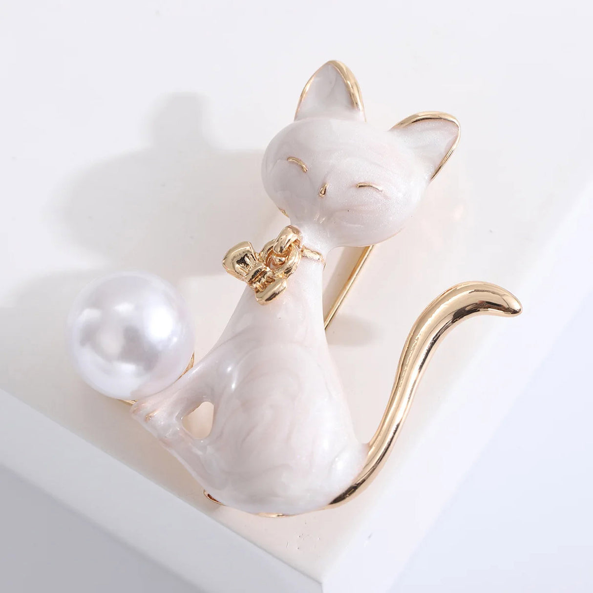 Cat brooch with white enamel body, gold-tone details, and white round bead, angled view on white background in studio.