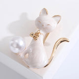 Cat brooch with white enamel body, gold-tone details, and white round bead, angled view on white background in studio.