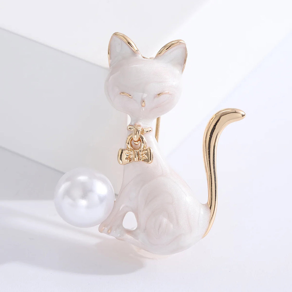 Cat brooch in white enamel with gold-tone tail and bow, holding a white round bead, upright view on white block.