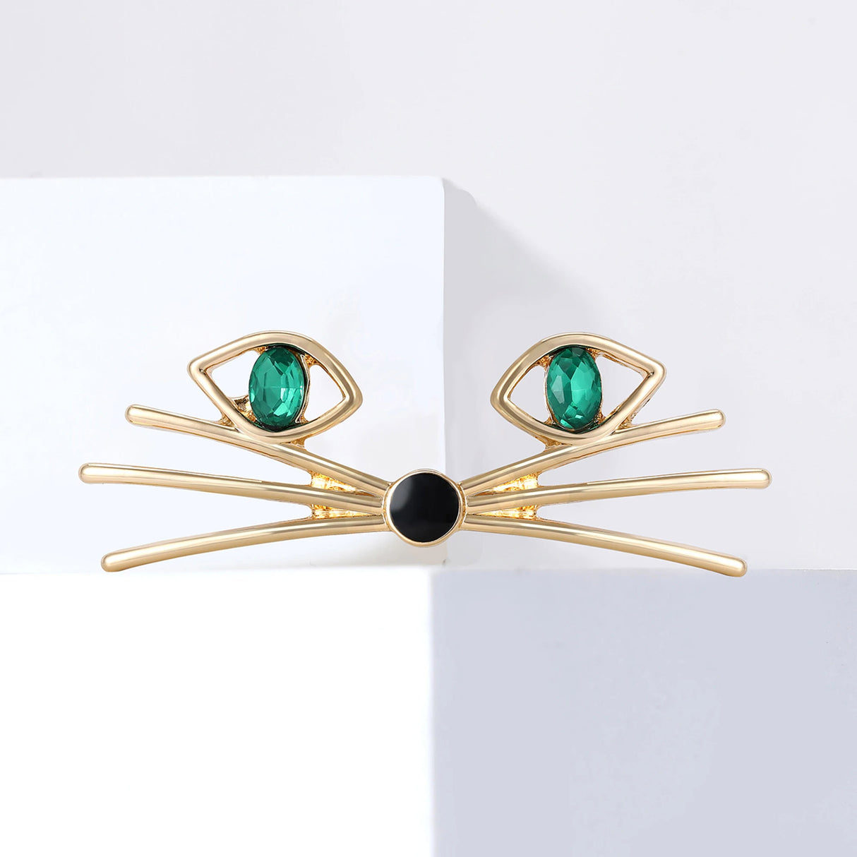 Cat face brooch in gold-tone with green oval stones for eyes and black round nose, displayed on white block.