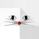 Cat face brooch in silver-tone metal with black oval stones for eyes and red round enamel nose, displayed on white block.