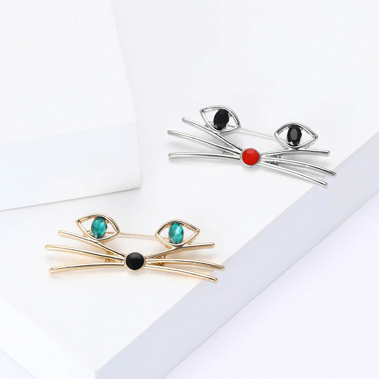Pair of cat face brooches in gold-tone with green stones and silver-tone with black stones, flat lay on white surface.