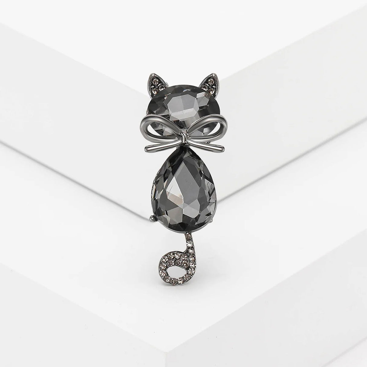Cat brooch with black faceted stones, dark metallic bow detail, and curled tail with small clear stones, upright pose on white studio background.