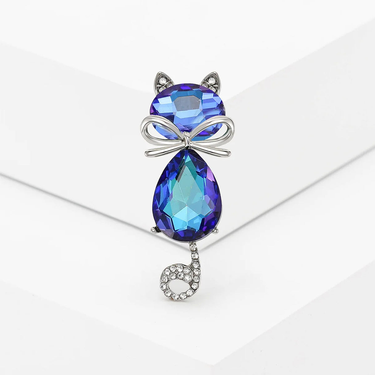Cat brooch with iridescent blue and purple faceted stones, silver-tone bow detail, and curled tail with clear stones, upright pose on white studio backdrop.