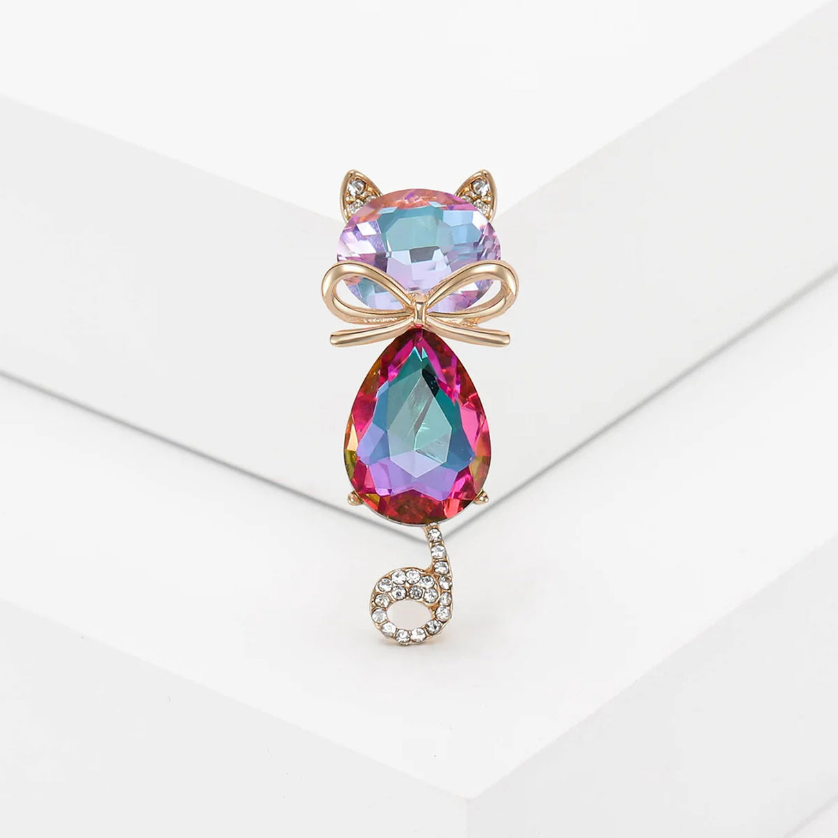 Cat brooch with iridescent pink faceted stones, gold-tone bow detail, and curled tail with clear stones, upright pose on white studio backdrop.
