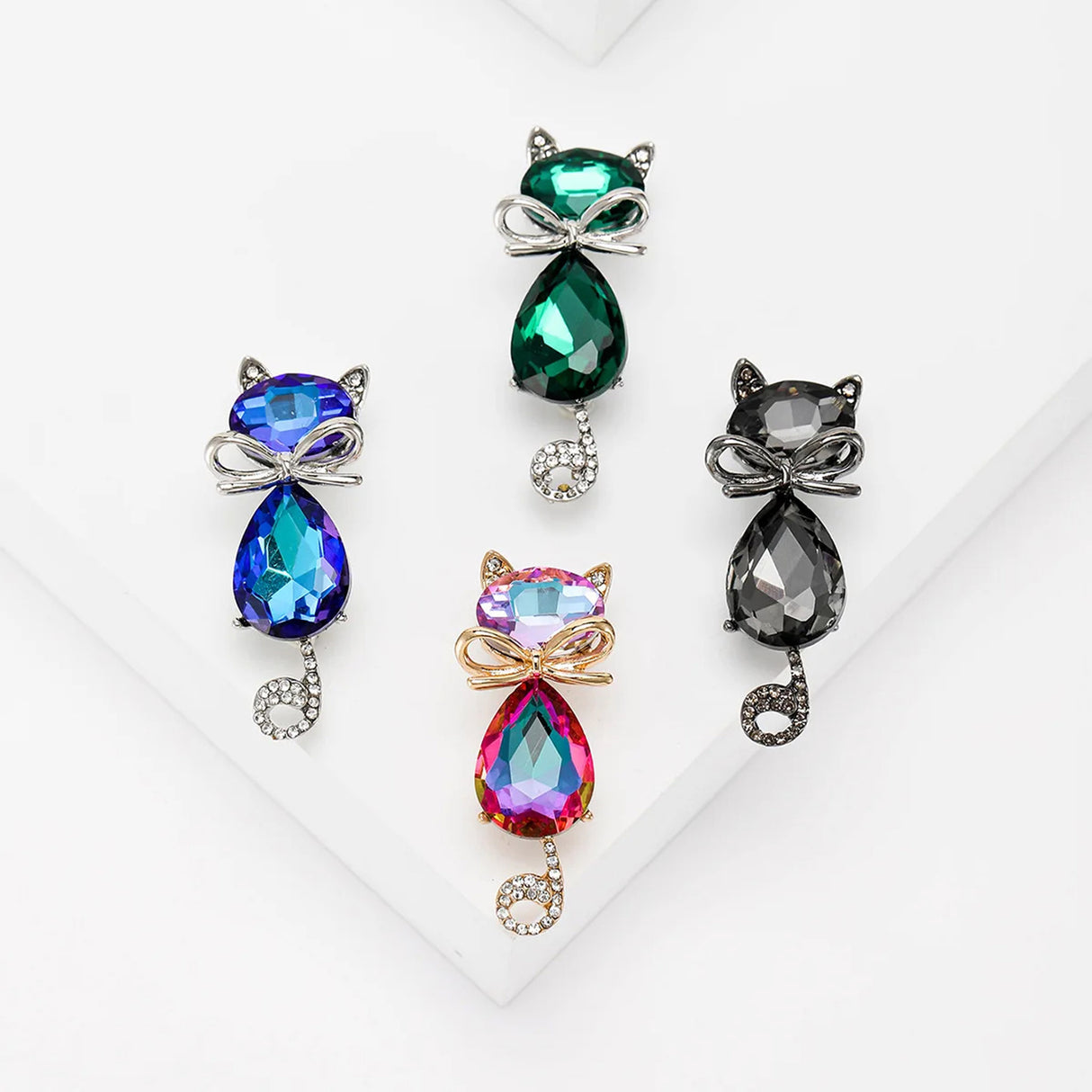 Set of four cat brooches in black, green, blue, and pink tones with faceted stones and curled tails, flat lay on white surface.
