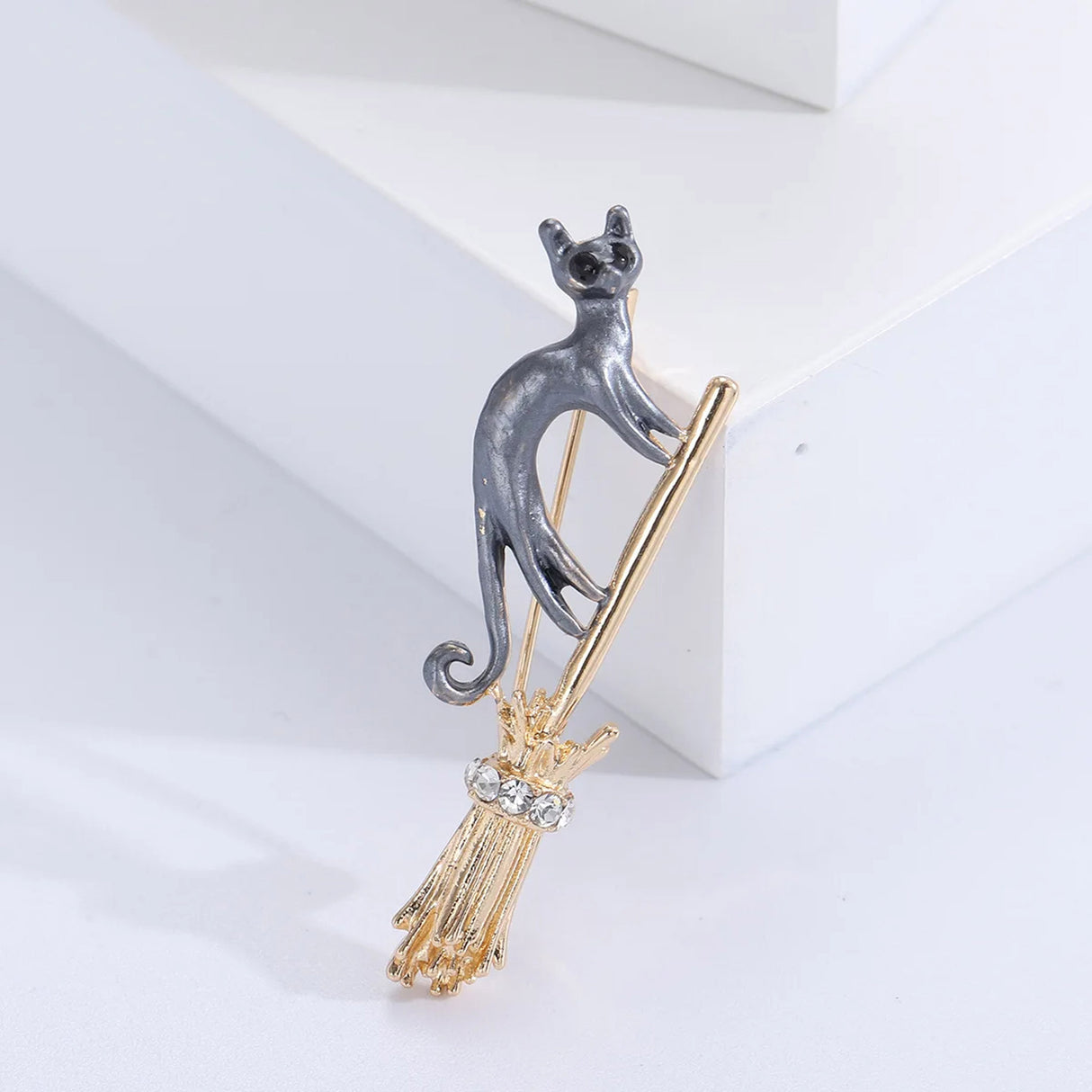 Cat brooch featuring gray-tone body standing on gold-tone broomstick with clustered clear stones, angled studio view on white block.