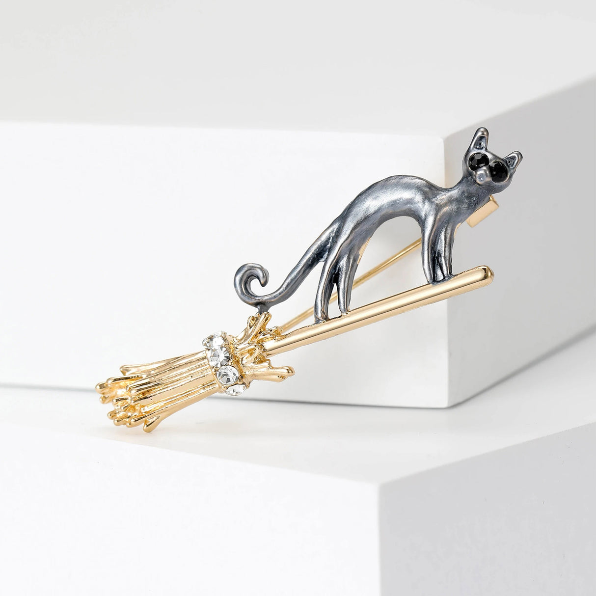 Cat brooch with dark gray body perched on gold-tone broomstick, clear faceted stones, front studio view.