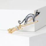 Cat brooch with dark gray body perched on gold-tone broomstick, clear faceted stones, front studio view.