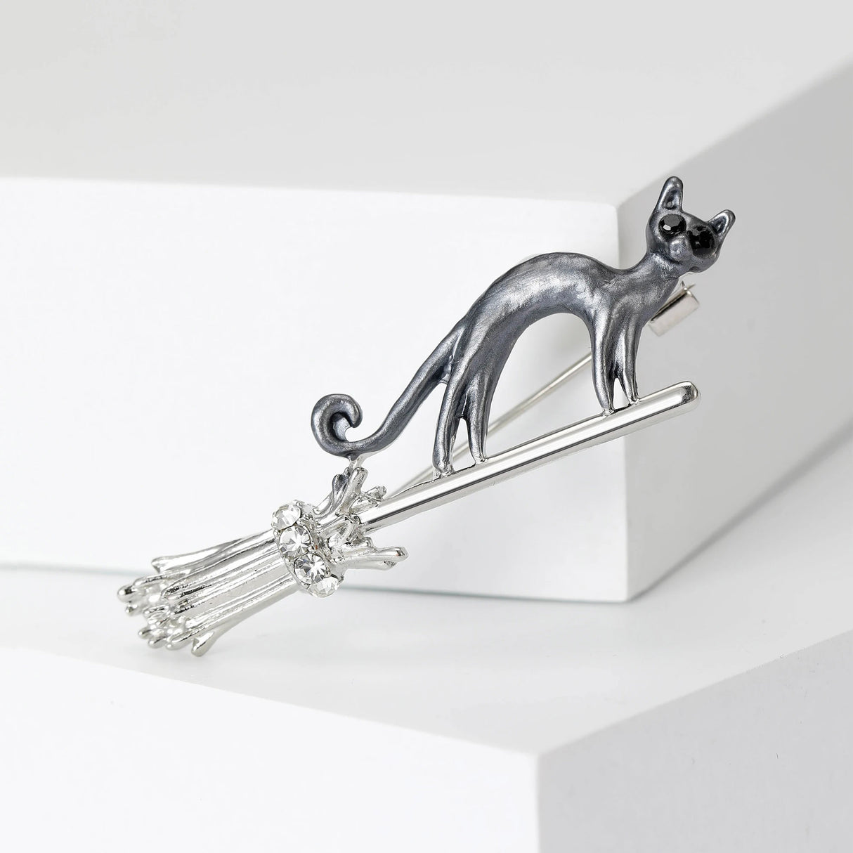 Cat brooch with gray-tone body positioned on silver-tone broomstick with faceted stones, front studio view on white block.