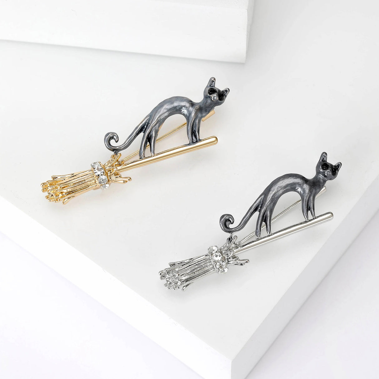 Pair of cat brooches with black bodies, one on gold-tone and one on silver-tone broomsticks, with clear stones, flat lay on white.