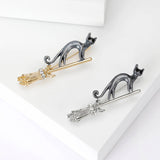 Pair of cat brooches with black bodies, one on gold-tone and one on silver-tone broomsticks, with clear stones, flat lay on white.