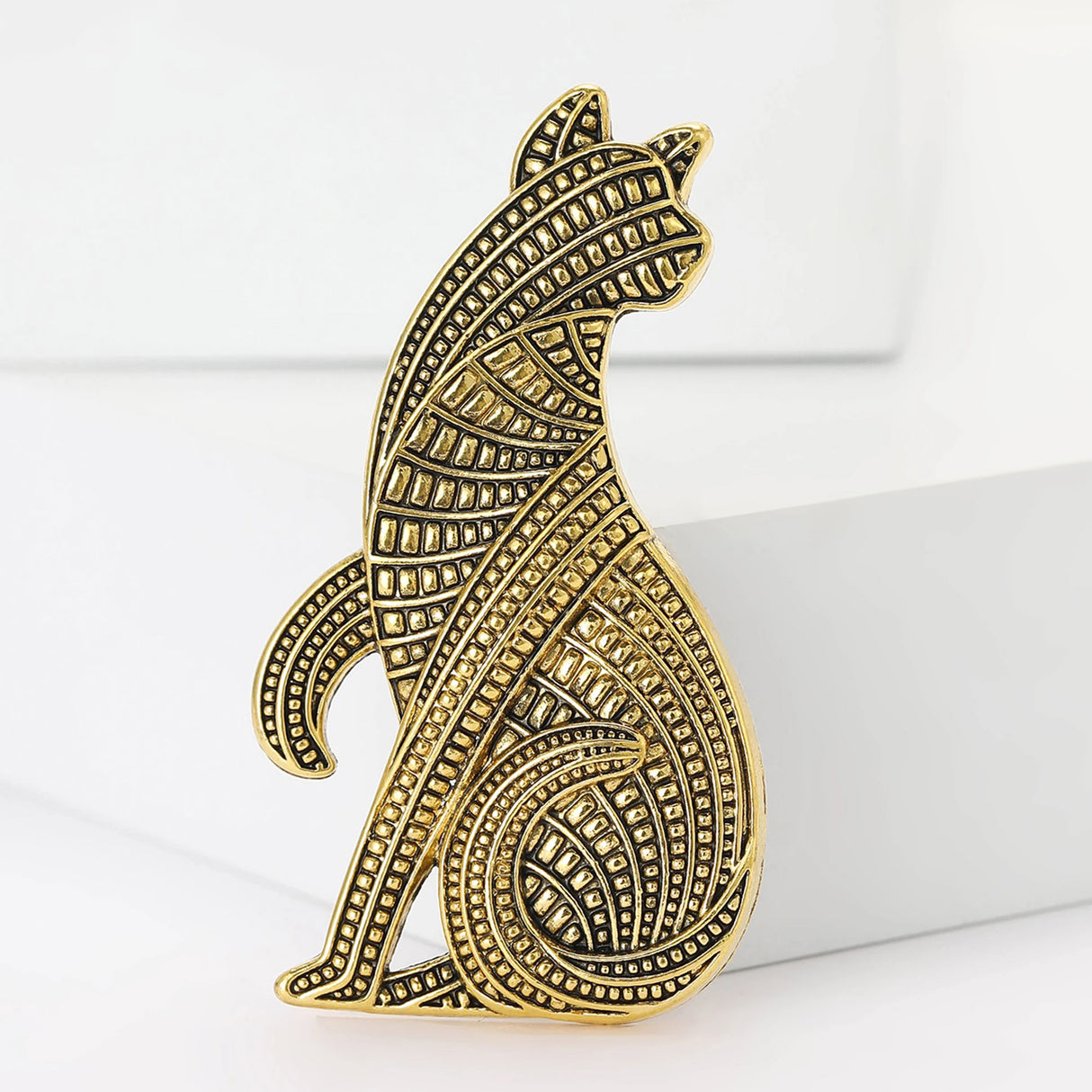 Cat brooch with gold-tone metal finish and embossed pattern, standing view on white block.