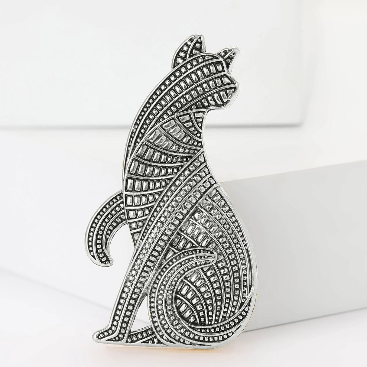 Cat brooch with silver-tone finish and patterned grid texture, upright pose on white platform in studio.