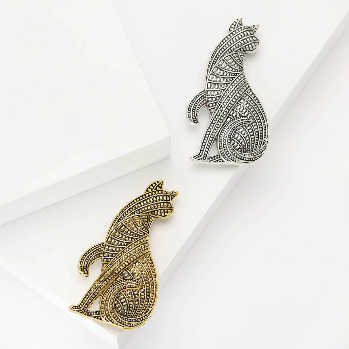 Pair of cat brooches in gold-tone and silver-tone metal finishes with geometric textures, flat lay on white surface.