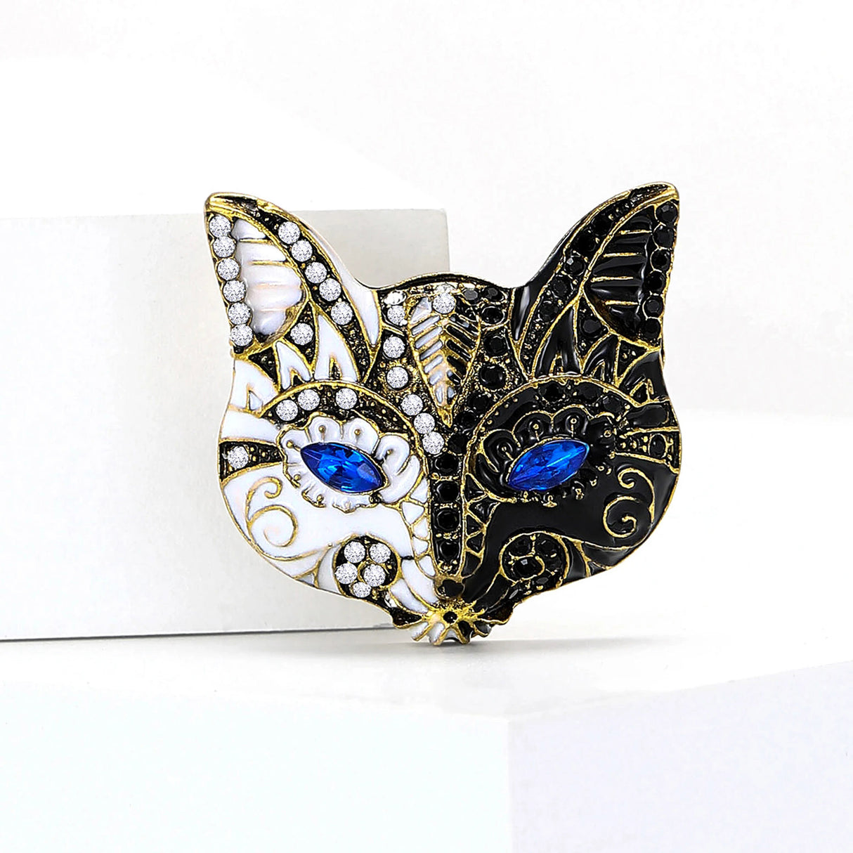 Cat mask brooch with black and white enamel design, gold-tone outline, blue marquise stone eyes, and round accents, upright view on white block.