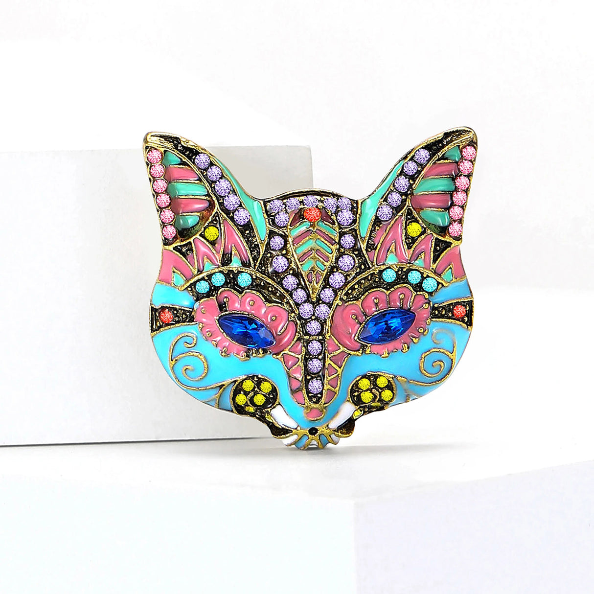 Cat mask brooch with turquoise blue and pink enamel, multicolor stones, and blue marquise eyes, upright view on white platform.