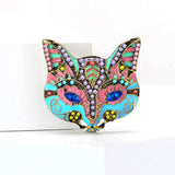 Cat mask brooch with turquoise blue and pink enamel, multicolor stones, and blue marquise eyes, upright view on white platform.