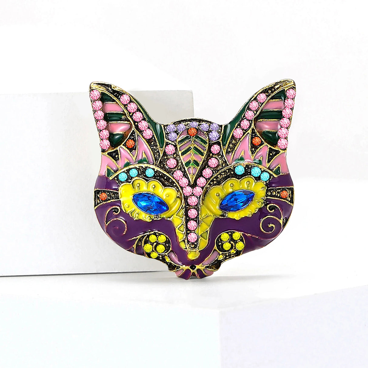 Cat mask brooch with purple and yellow enamel, multicolor stones, and blue marquise eyes, upright view on white platform.