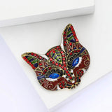 Cat mask brooch with red and brown enamel, blue marquise eyes, and red and green decorative details, angled view on white surface.