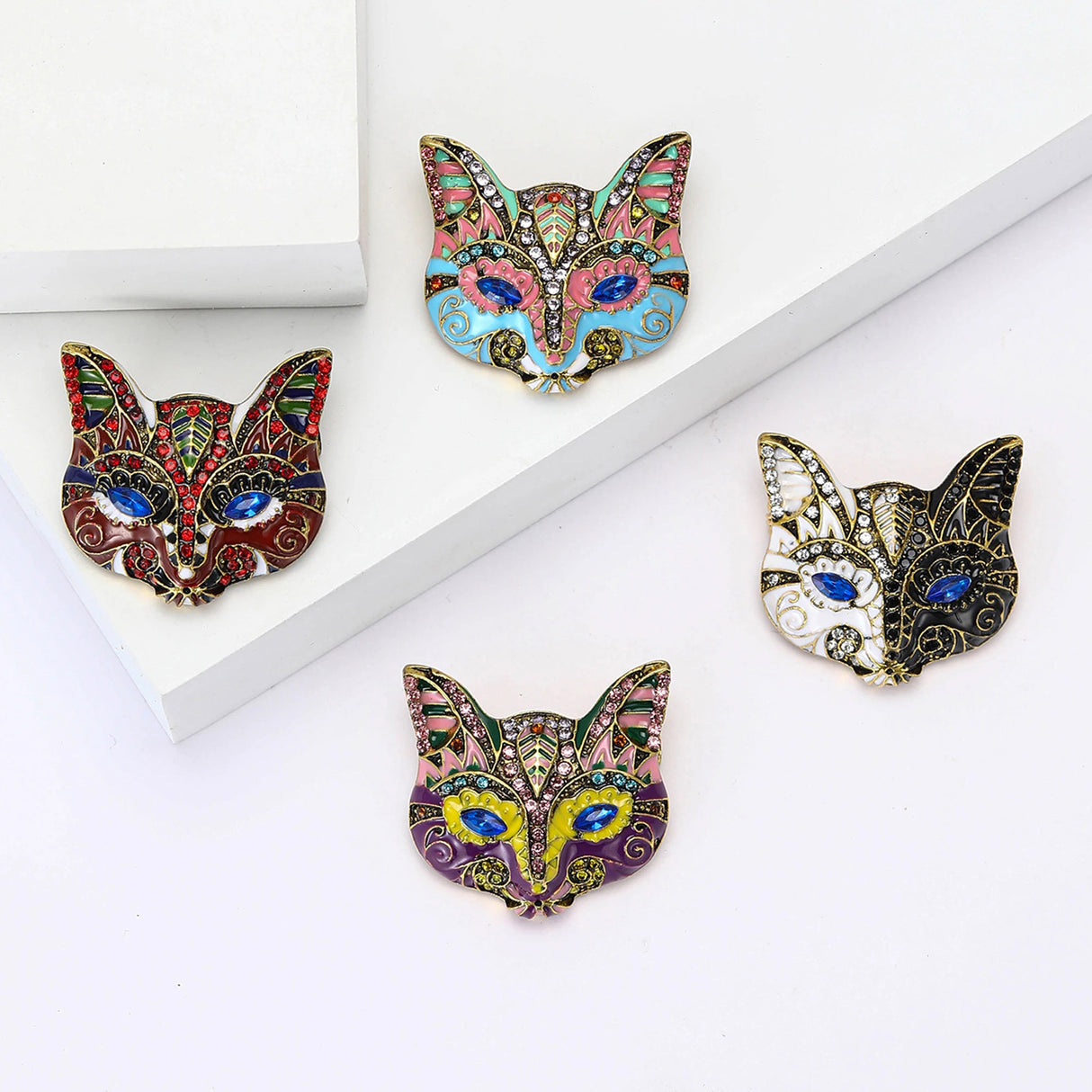 Set of four cat mask brooches in multicolor enamel with blue marquise stones for eyes and decorative accents, flat lay on white surface.