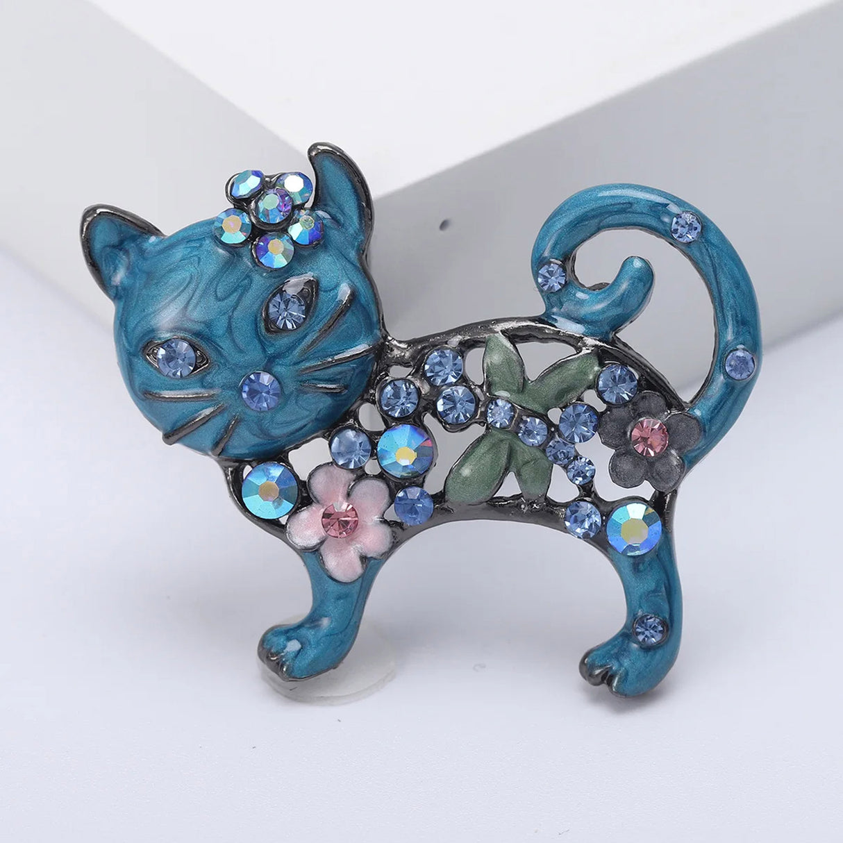 Cat brooch with blue enamel finish, blue and pink stones, floral accents, and green leaf detail, angled view.