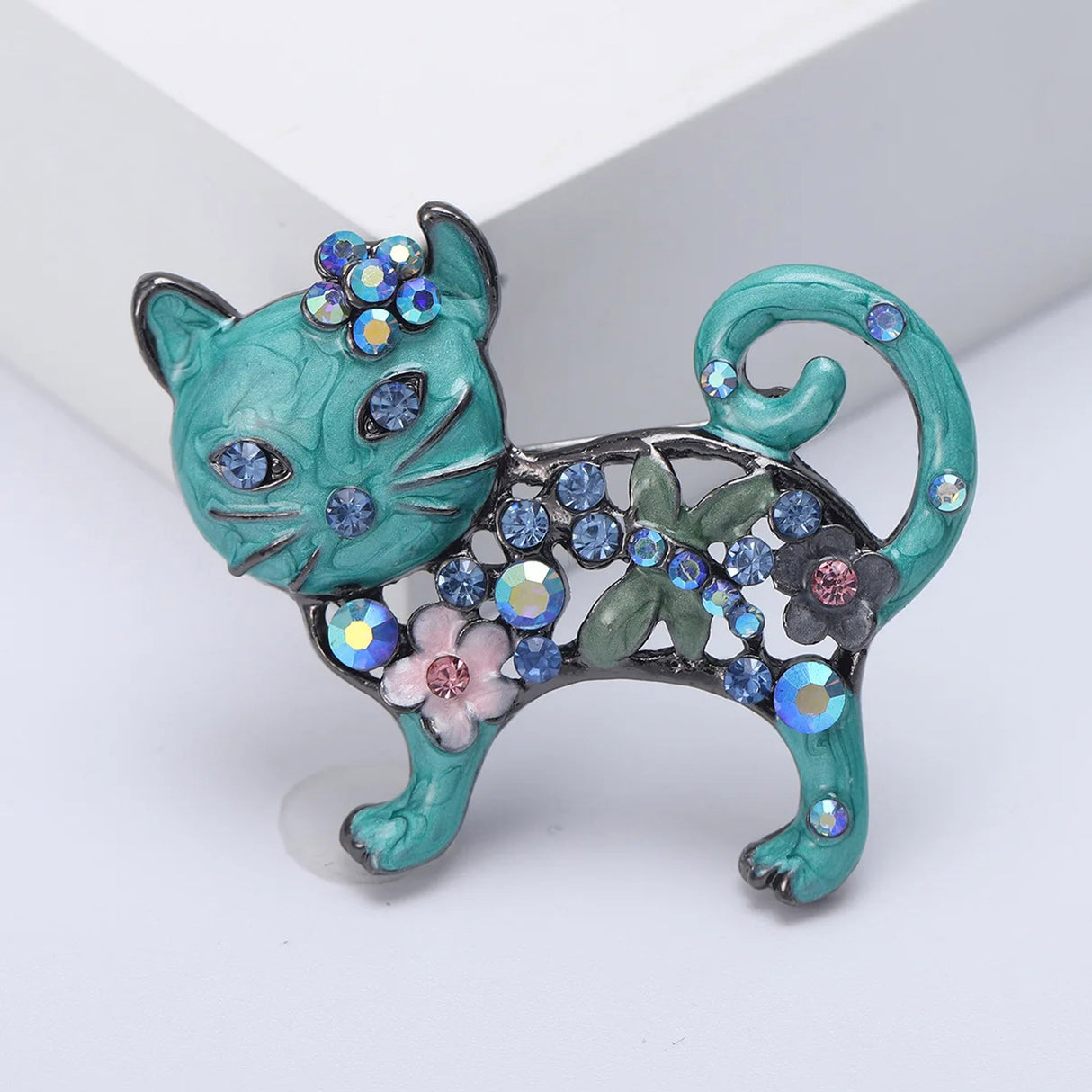 Cat brooch with teal-blue enamel, blue decorative stones, pink flower detail, and green leaf motif, upright studio pose.