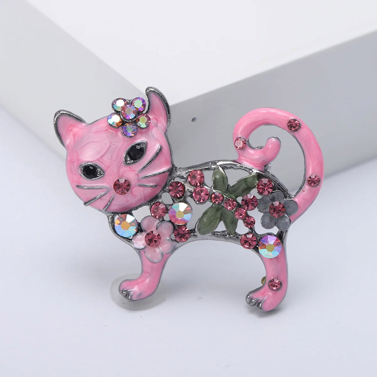 Cat brooch with pink enamel and round faceted stones, green leaf accent, and multicolor floral details, front view in studio.